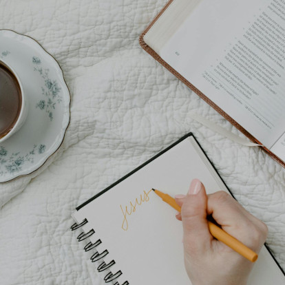 Mão escrevendo o nome Jesus em um caderno sobre mesa rústica com Bíblia aberta e xícara de café